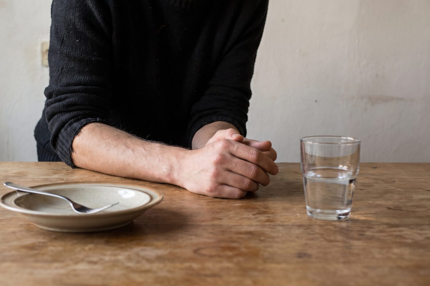 Iemand zit met de armen op een keukentafel bij een leeg bord en een glas water