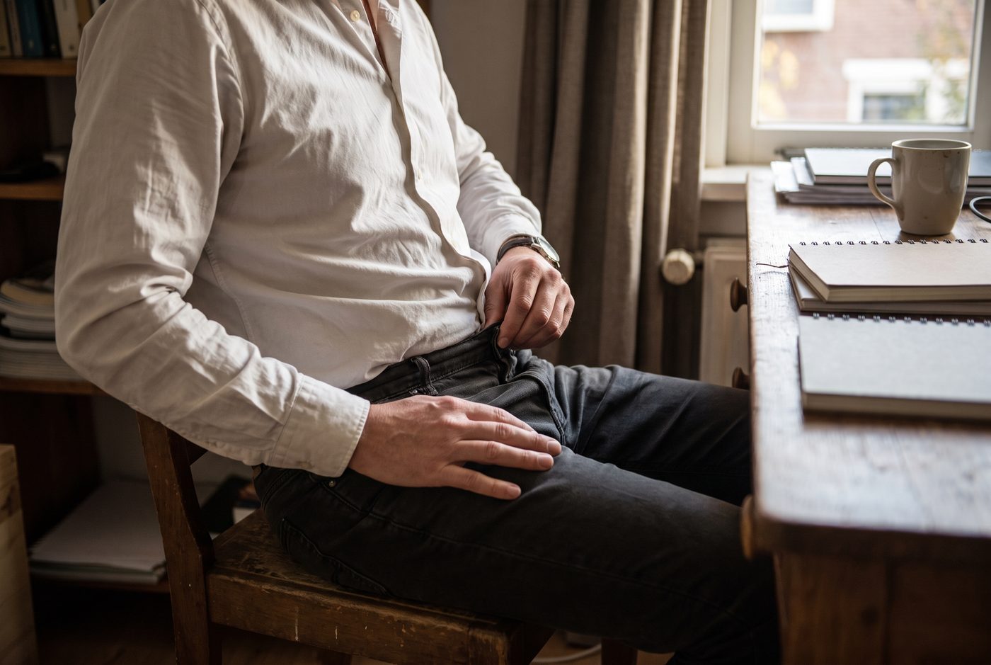 Iemand zit aan een bureau en maakt de bovenste knoop van de broek los, gezien vanaf borsthoogte