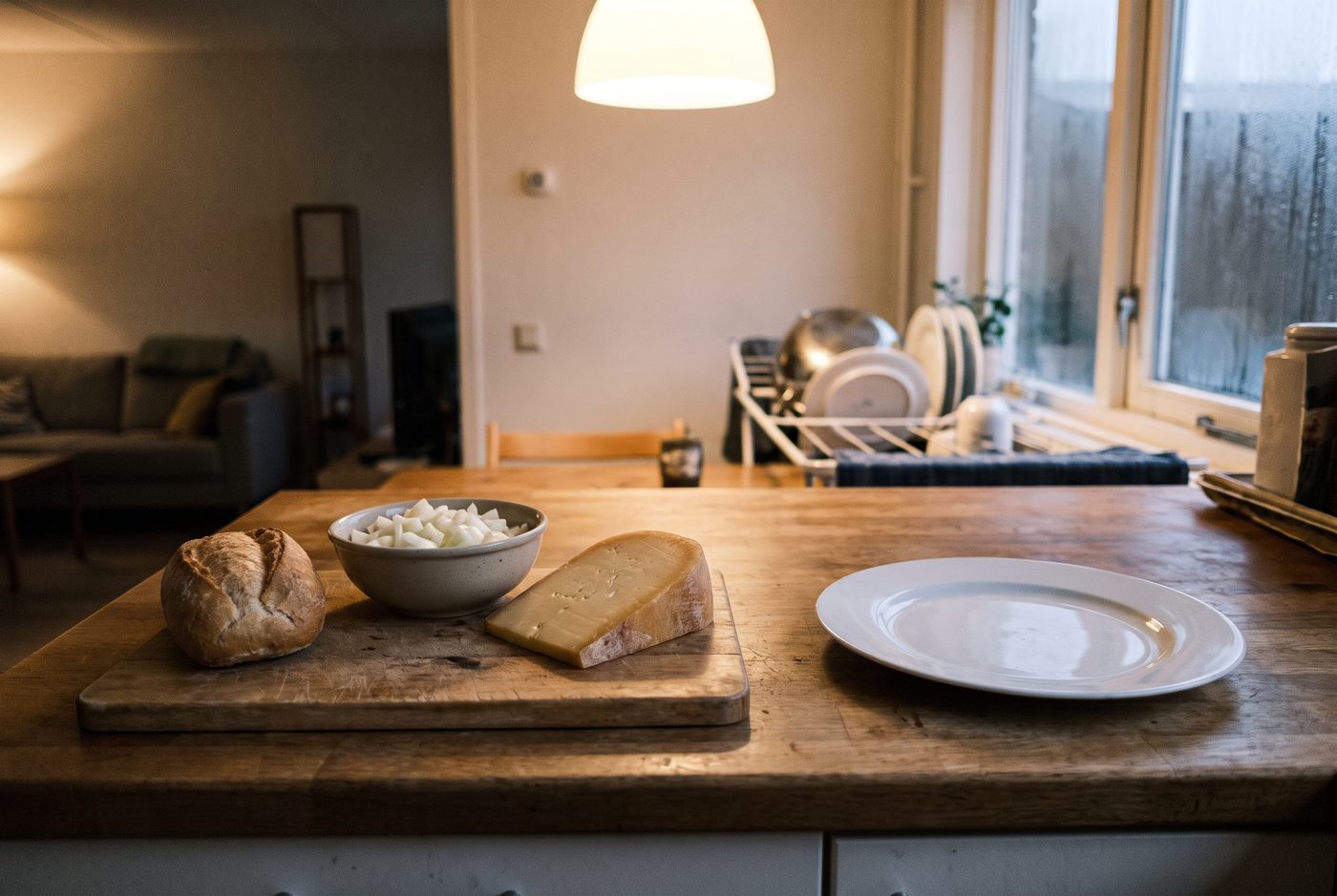 Een broodje, een schaaltje gesneden rauwe ui en een punt kaas op een snijplank naast een leeg bord