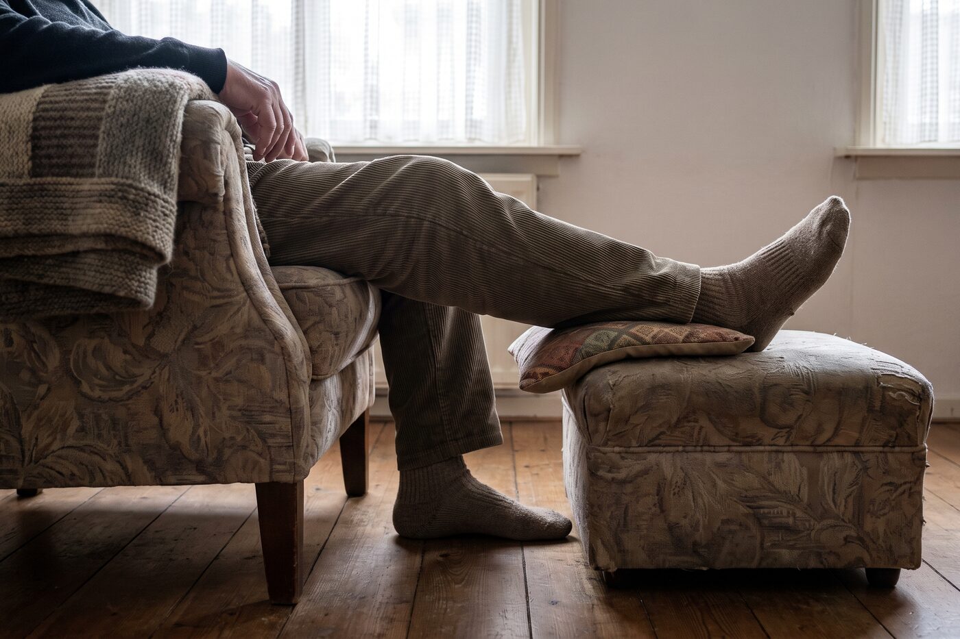 Iemand zit in een fauteuil met een been op een voetenbankje en een kussen onder de knie