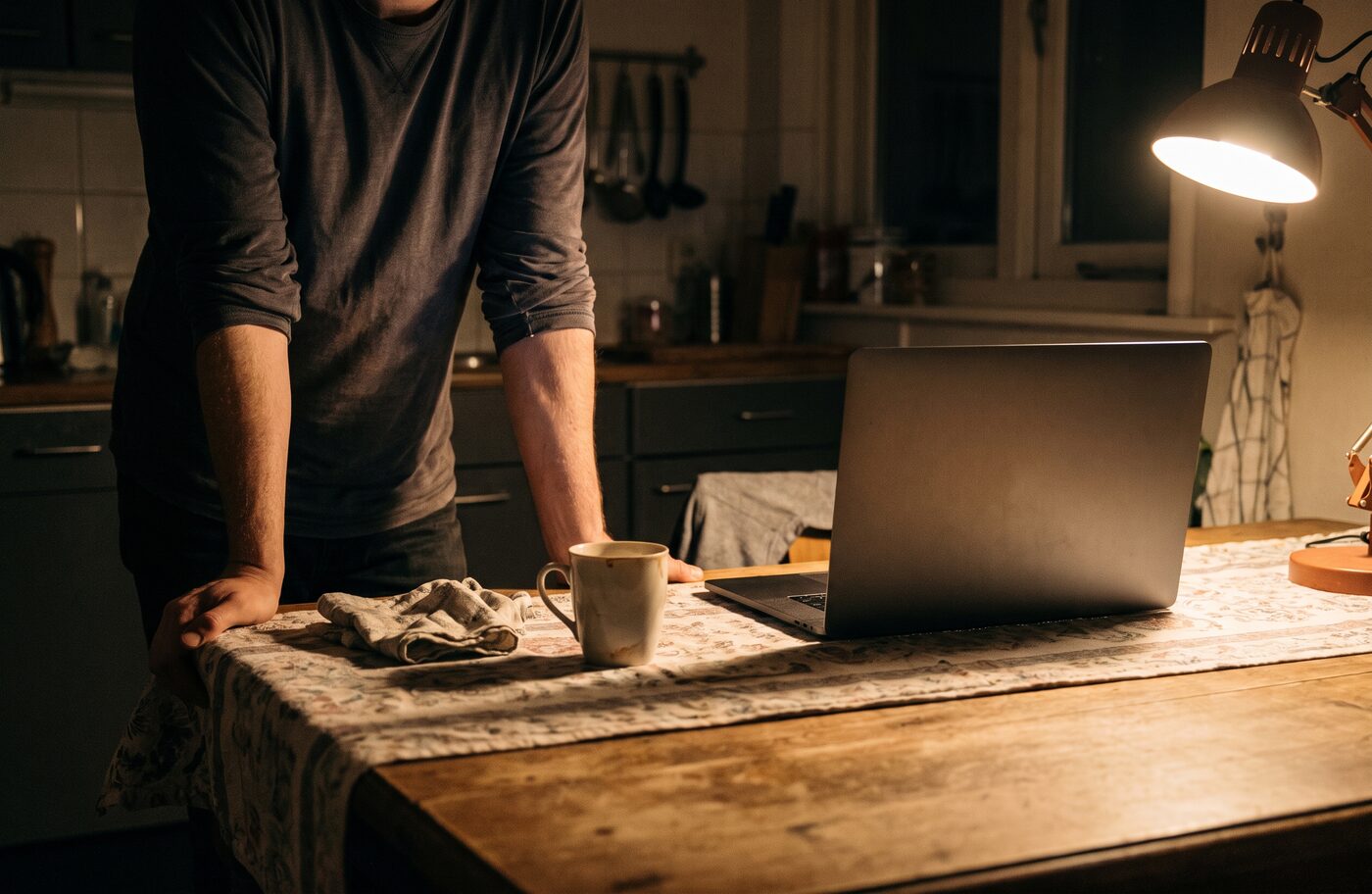 Iemand staat 's nachts aan de keukentafel bij een open laptop en een lamp