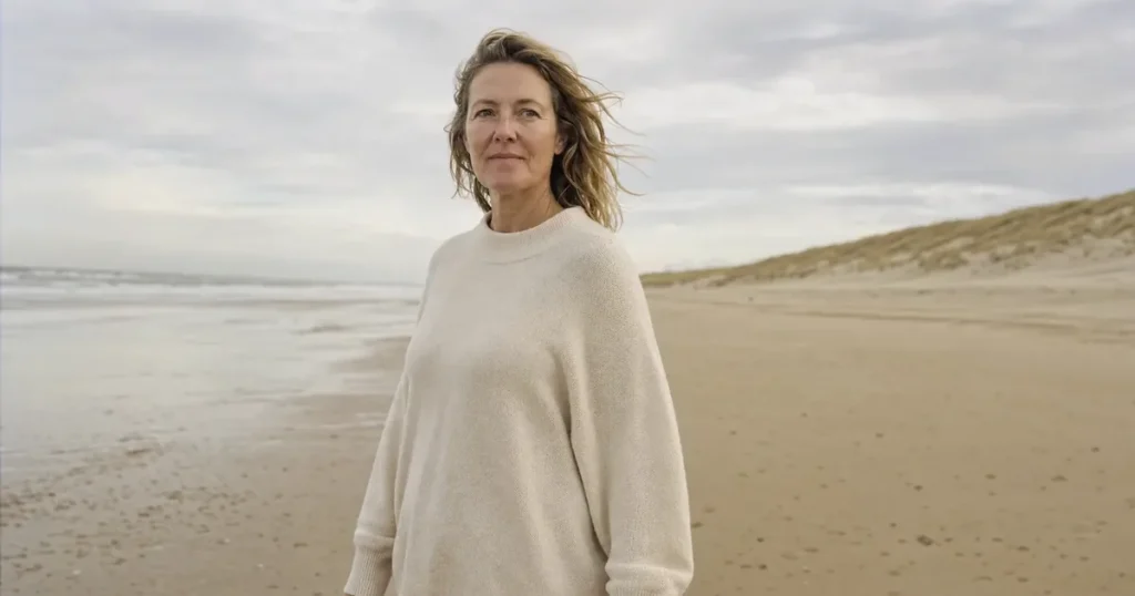 Een vrouw van rond de 50 met donkerblond haar in de wind en een ruime cremekleurige trui staat op een breed Noordzeestrand langs de duinen, bij een artikel over de buik na bevalling.