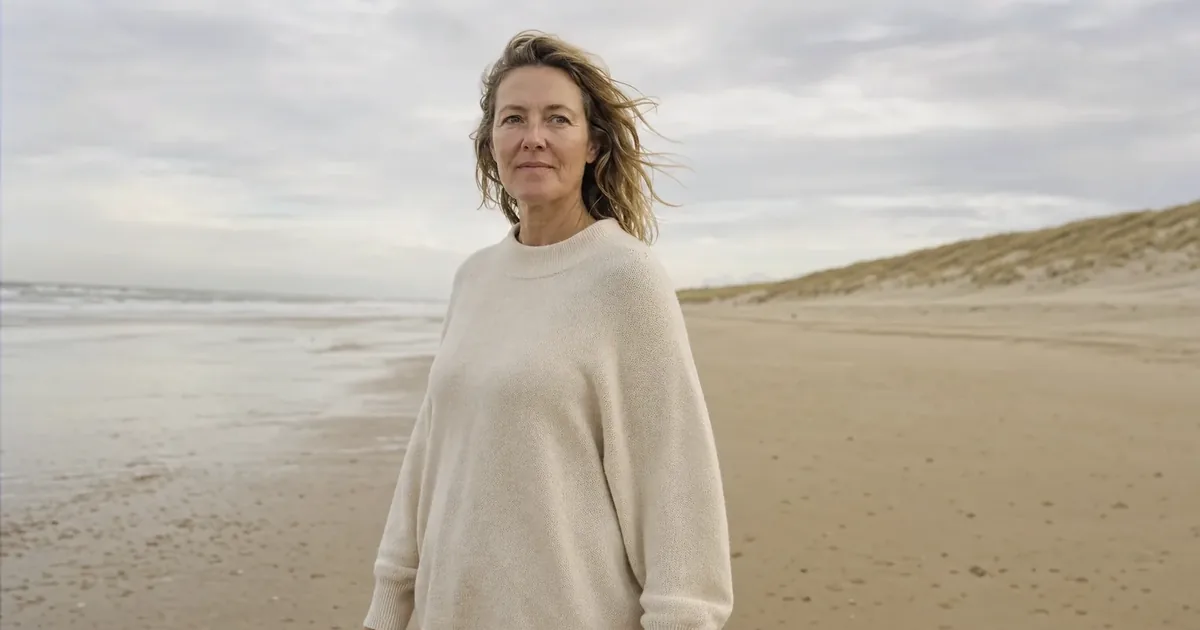 A woman of around 50 with dark blonde hair in the wind and a loose cream coloured jumper stands on a wide North Sea beach along the dunes, for an article about the belly after birth.