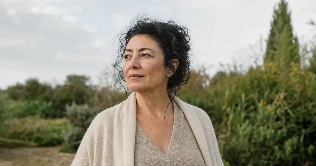 Beeld bij hartklachten vrouwen: portret van een vrouw van ongeveer 52 met donker krullend haar in een tuin, de blik rustig langs de camera.