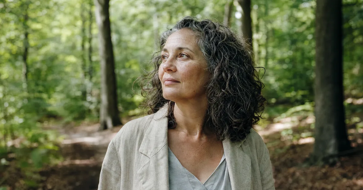 Portrait of a woman of around 48 with greying curly hair in a sunny beech wood, looking calmly to one side.