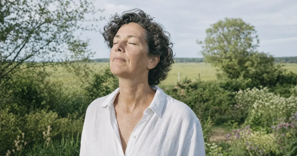 Een vrouw van rond de 50 met kort donker krullend haar en een wit linnen overhemd staat met gesloten ogen en het gezicht naar de zon in een groene tuin aan de rand van een vlak weiland, een rustig moment dat past bij je beter voelen na de overgang.
