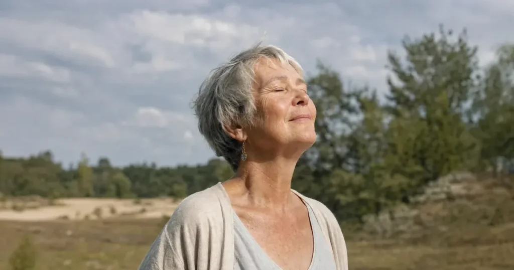 Een vrouw van in de 60 met kort grijs haar houdt aan de rand van een bos de ogen even dicht in de zon, een korte pauze tijdens de middagdip.