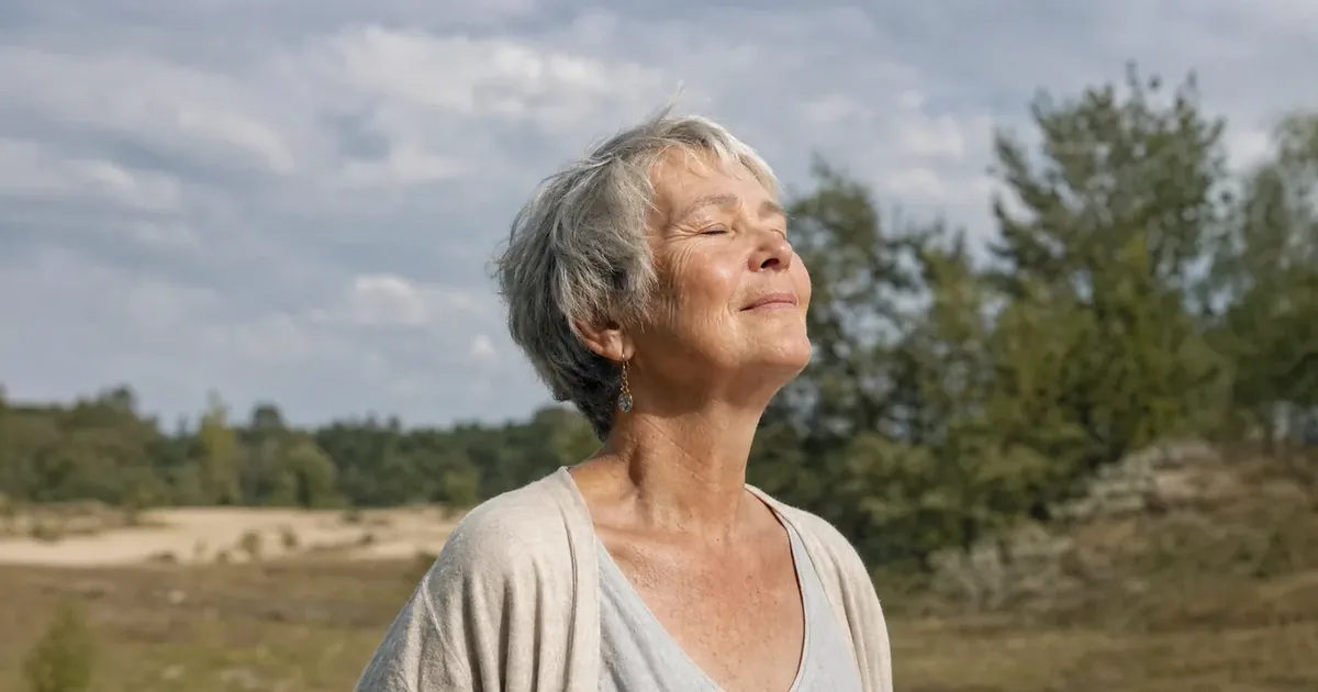 A woman of about 60 with short grey hair at the edge of a wood, eyes closed for a moment in the sun, a short break during the afternoon dip.