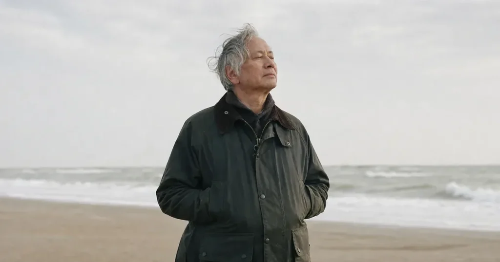 Een man van in de 60 met grijs haar staat met gesloten ogen in de zeewind op het strand, rustig, ook op dagen met een somber gevoel.