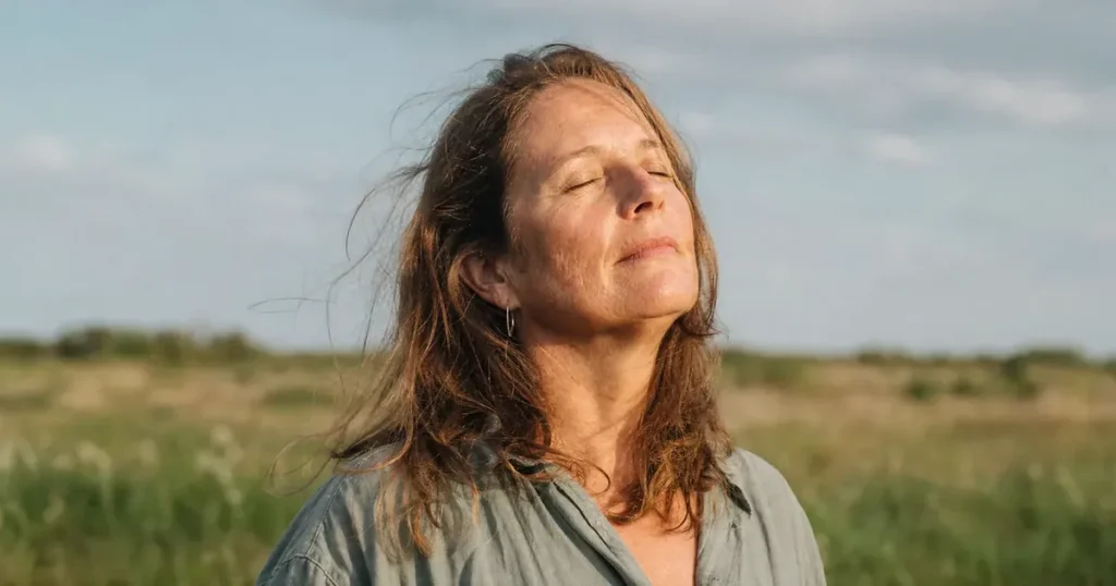 Een vrouw van rond de 50 staat met de ogen dicht en het gezicht naar de lage zon in een grasland, een moment rust bij stress symptomen.