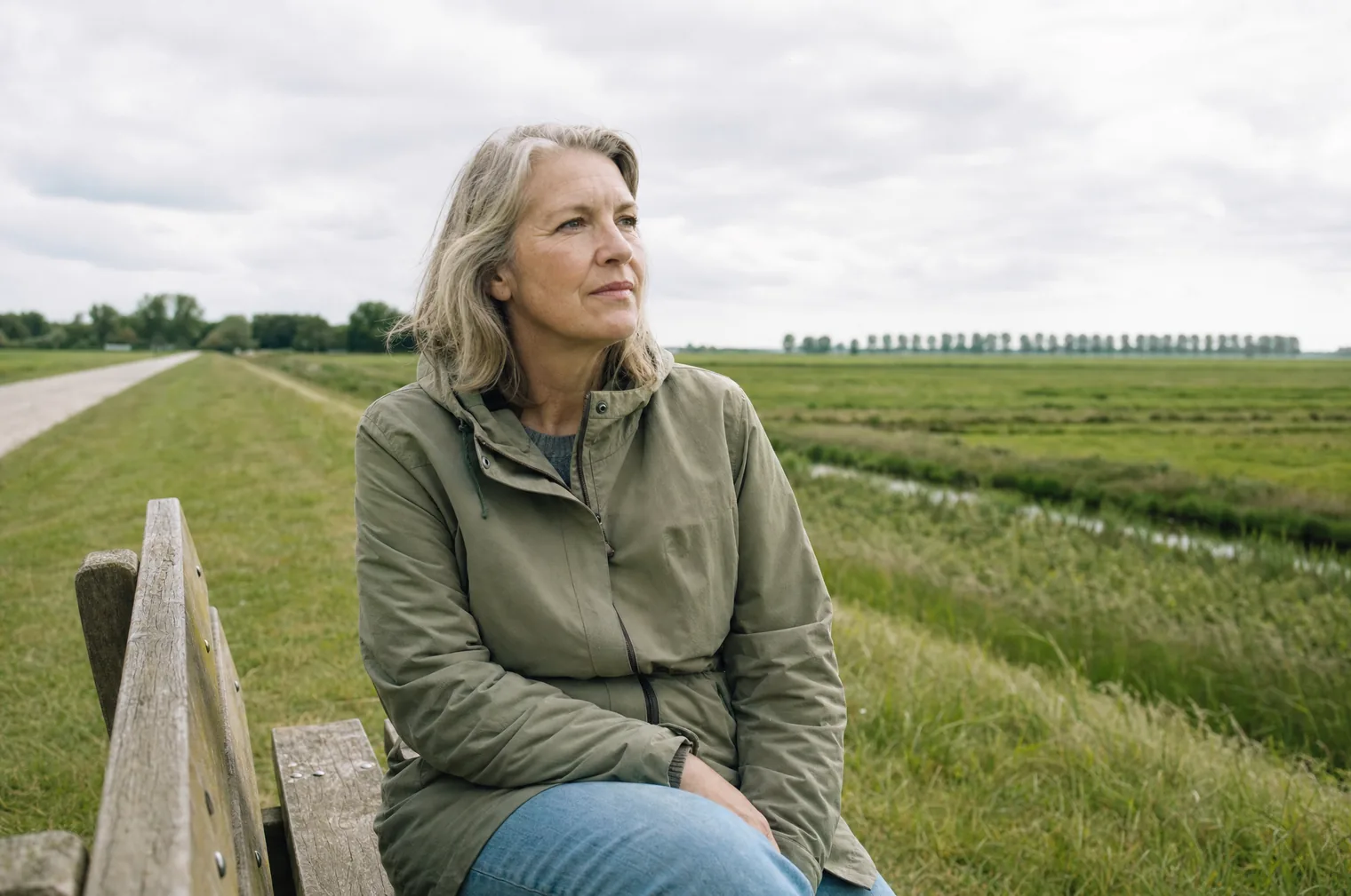 Eine Frau um die 55 mit graublondem Haar und einer olivgrünen Jacke sitzt auf einer Holzbank auf einem Deich und blickt über einen flachen Polder mit einem Graben und einer Reihe Pappeln.