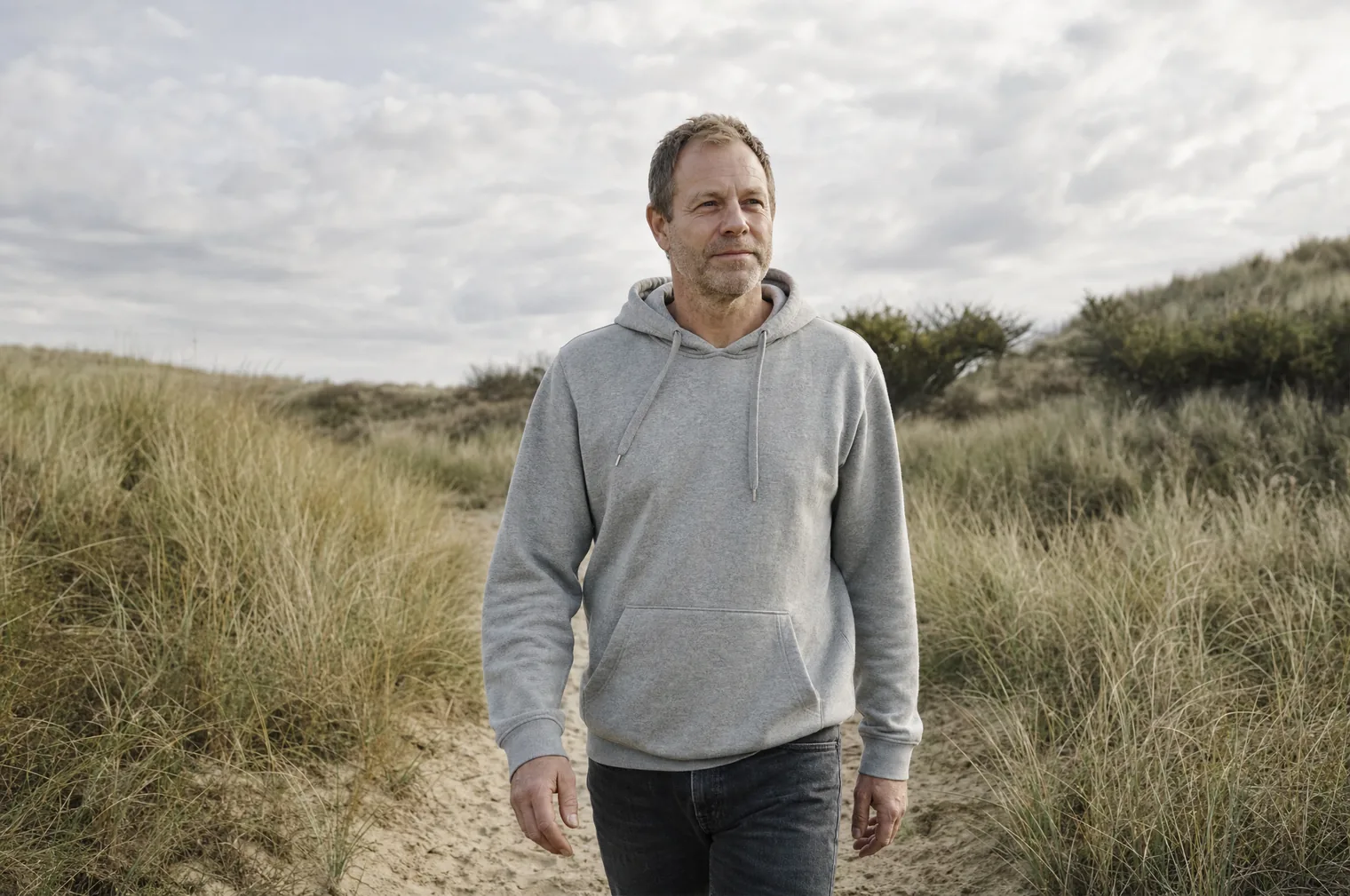 Ein Mann um die 50 mit kurzen hellbraunen Haaren und Bartstoppeln, in einem grauen Hoodie, geht auf einem schmalen Sandweg durch die Dünen mit Strandhafer.
