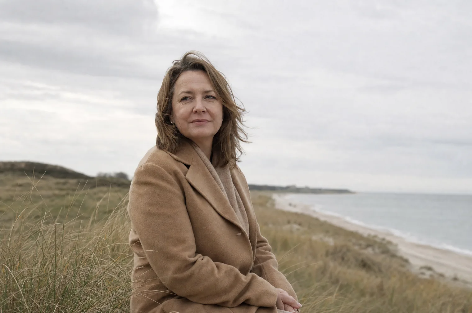 Eine Frau in den 50ern mit halblangen braunen Haaren und einem kamelfarbenen Mantel sitzt zwischen dem Strandhafer oben auf einer Düne, im Hintergrund ein langer Strand und das Meer.