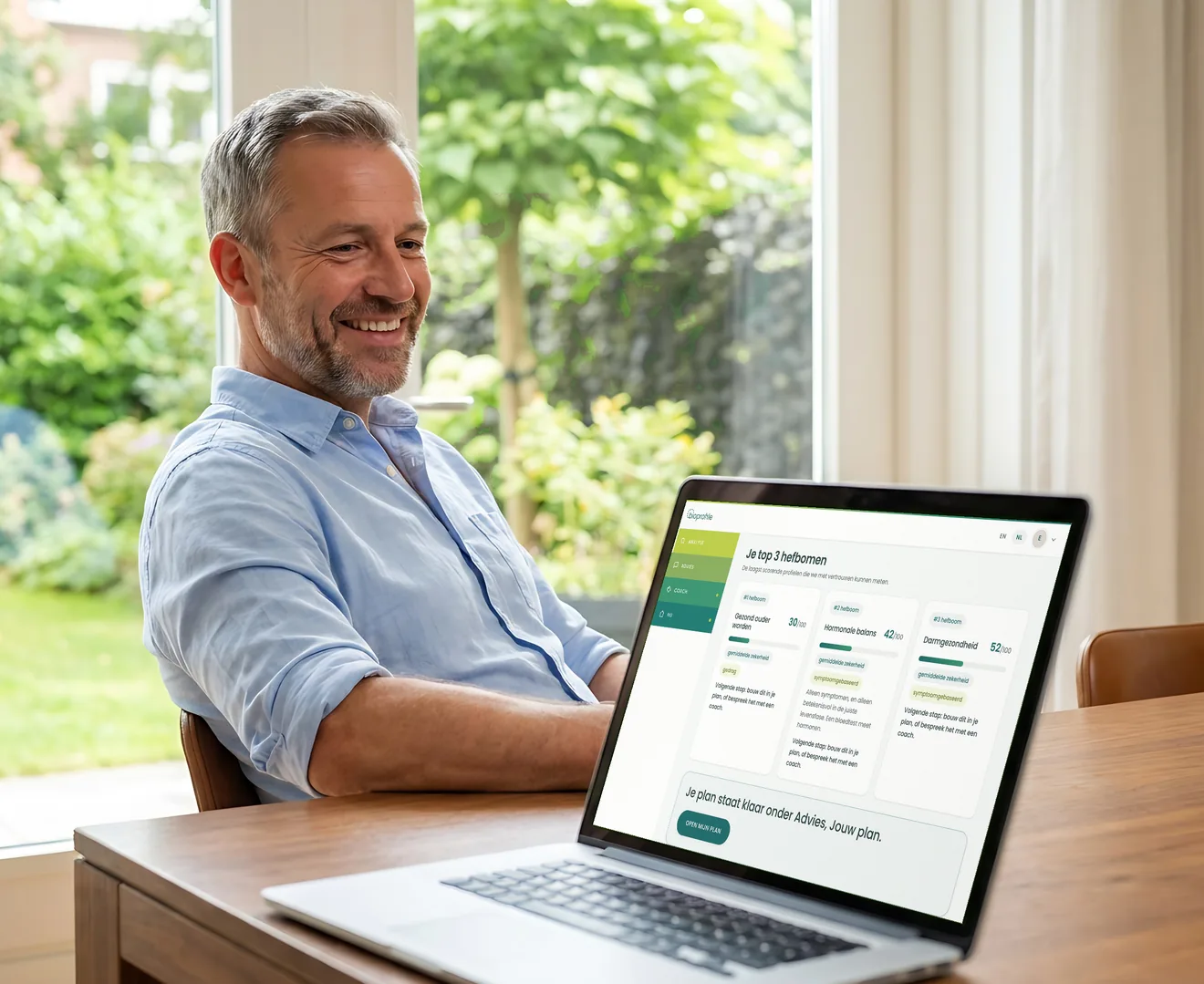 Ein Mann Anfang fünfzig in einem hellblauen Hemd sitzt an einem hölzernen Esstisch an einem Fenster mit Blick auf den Garten und schaut lächelnd auf den Laptop, mit den Analyseergebnissen der BioProfile-App auf dem Bildschirm: die drei größten Hebel mit ihren Scores.