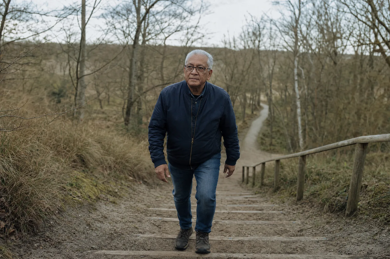 Ein Mann in den 60ern mit grauem Haar und Brille, in einer dunkelblauen Jacke, geht ruhig eine Holztreppe an einem Geländer hinauf, in einem Dünenwald mit Birken.