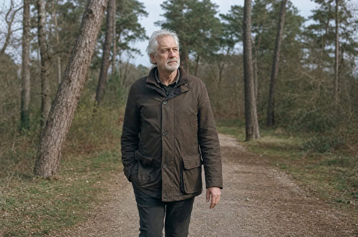 Ein Mann in den 60ern mit weißem Haar und kurzem weißem Bart, in einer braunen Feldjacke, geht über einen Kiesweg zwischen hohen Kiefern, eine Hand in der Hosentasche.