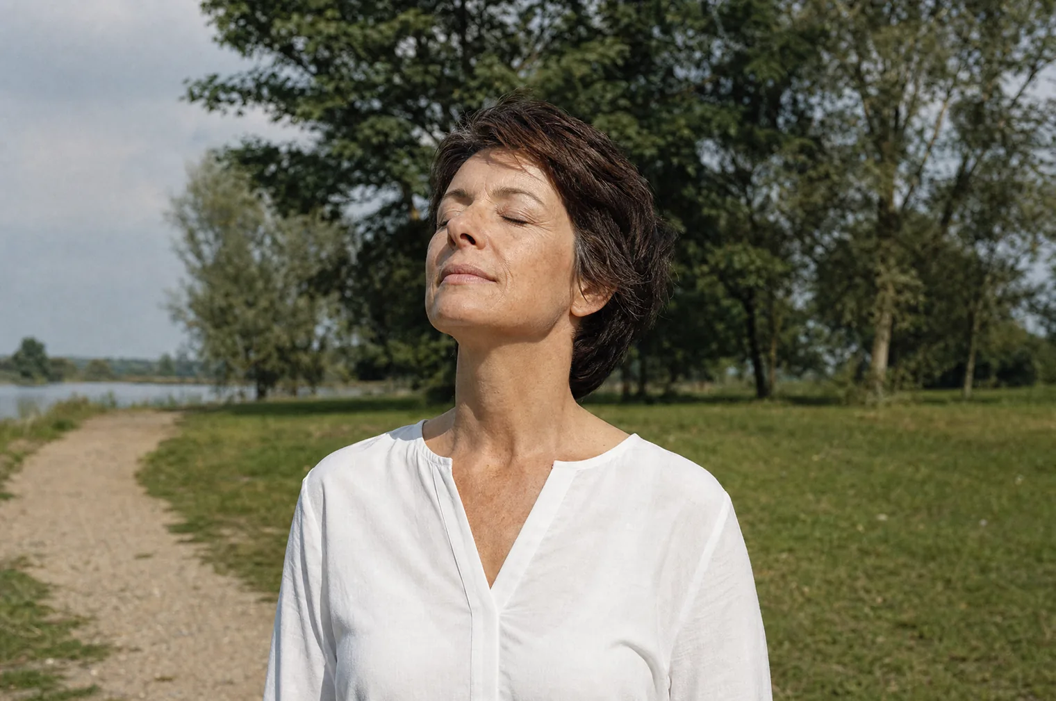 A woman of around 50 with short dark brown hair and a white blouse stands with eyes closed and face turned to the sun in a park by the water.