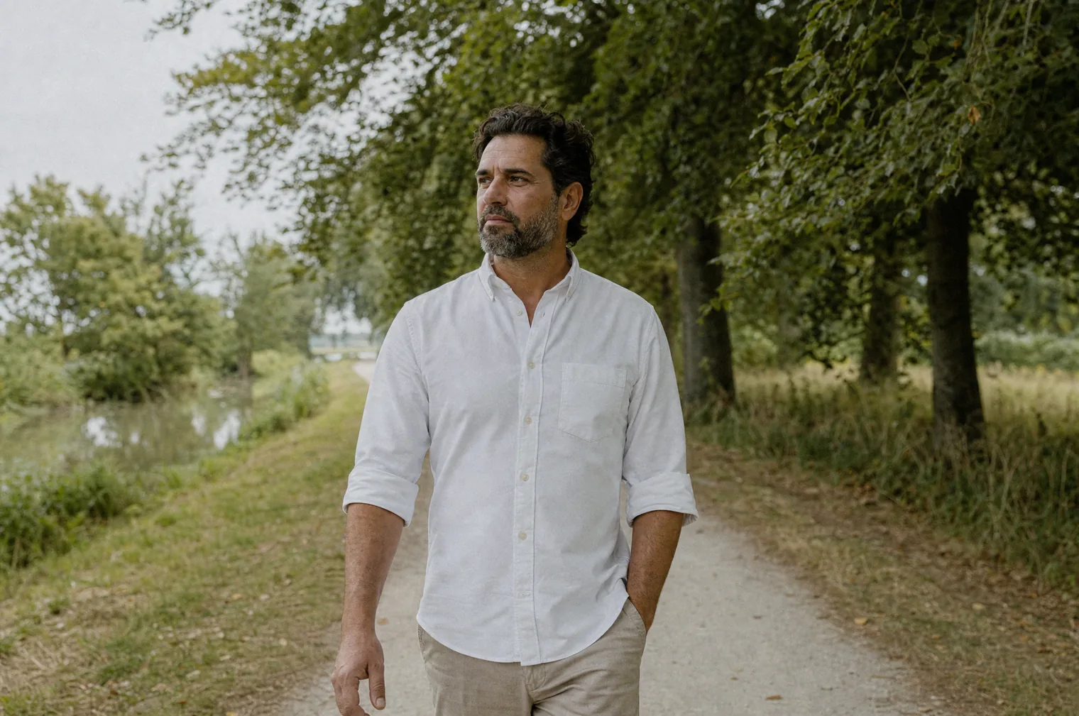 Un hombre de unos 40 años con el pelo oscuro y la barba canosa, con camisa blanca, camina por un sendero bajo una hilera de árboles junto a un canal.