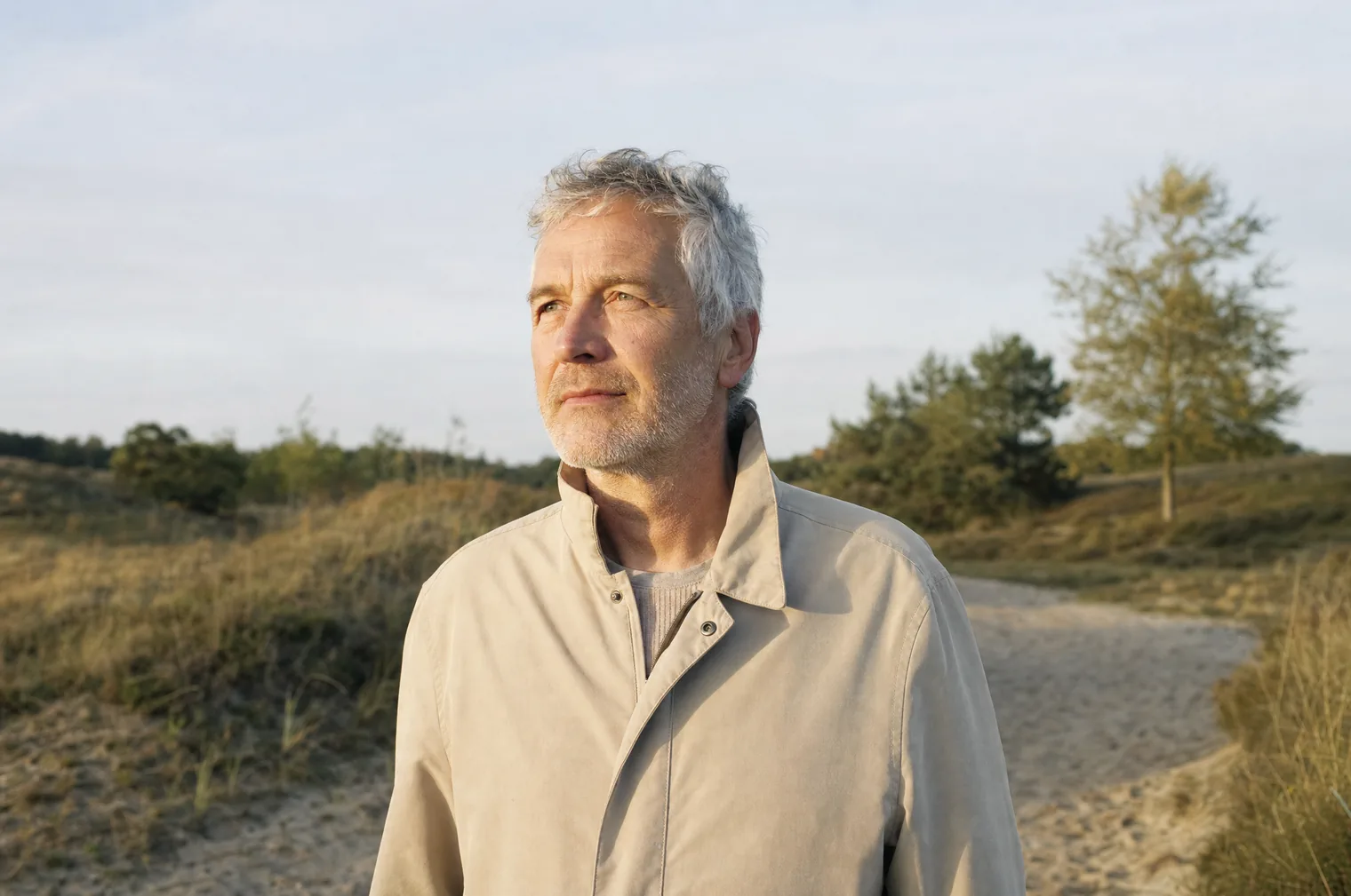 A man in their 50s with grey hair and a light jacket stands in the early morning sun on a sandy path through the dunes.