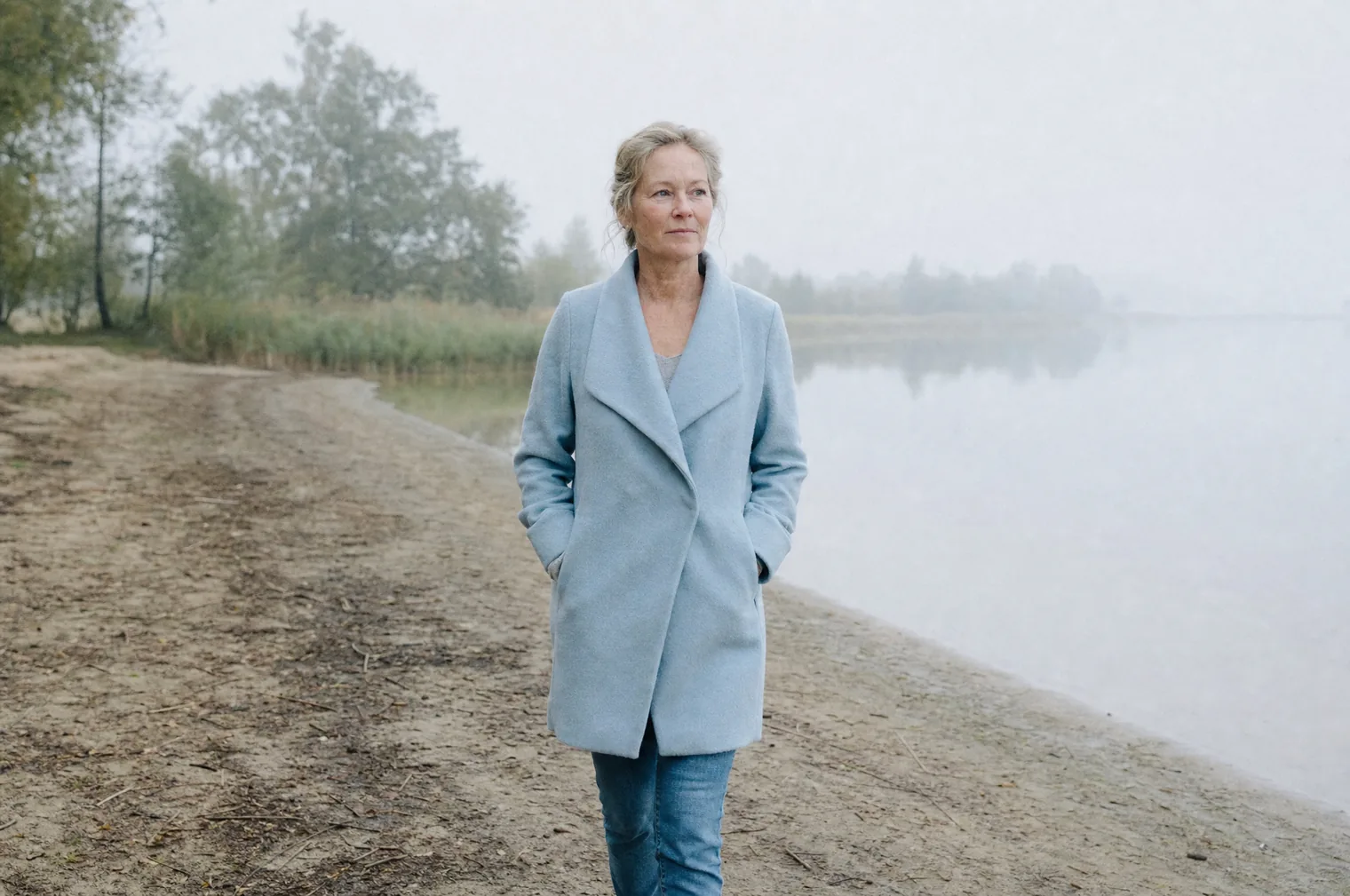 A woman in their 50s with blonde hair pinned up and a light blue jacket walks with hands in pockets along the shore of a lake in the morning mist.