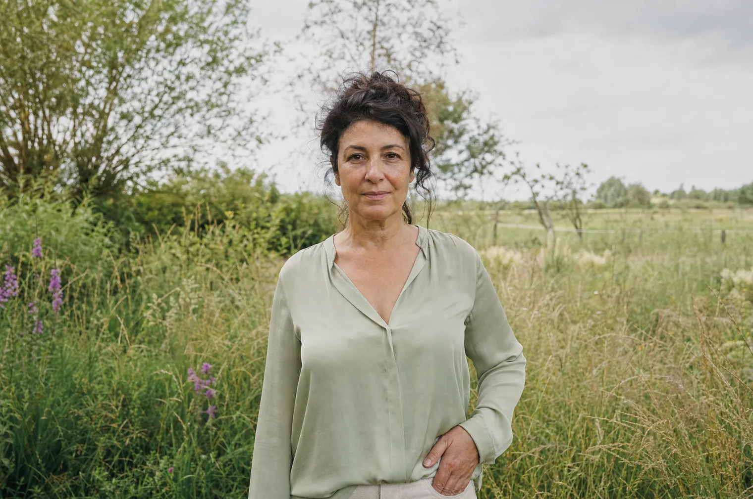 A woman of around 50 with dark curly hair pinned up and a light green blouse stands in tall grass beside a meadow, one hand on the hip.