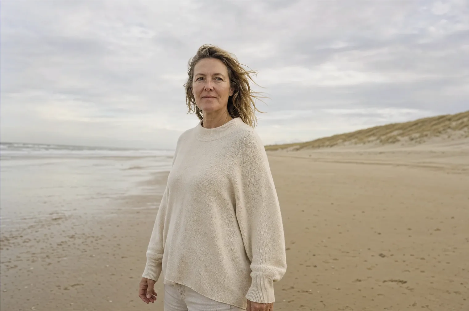 A woman of around 50 with dark blonde hair in the wind and a loose cream coloured jumper stands on a wide North Sea beach along the dunes.