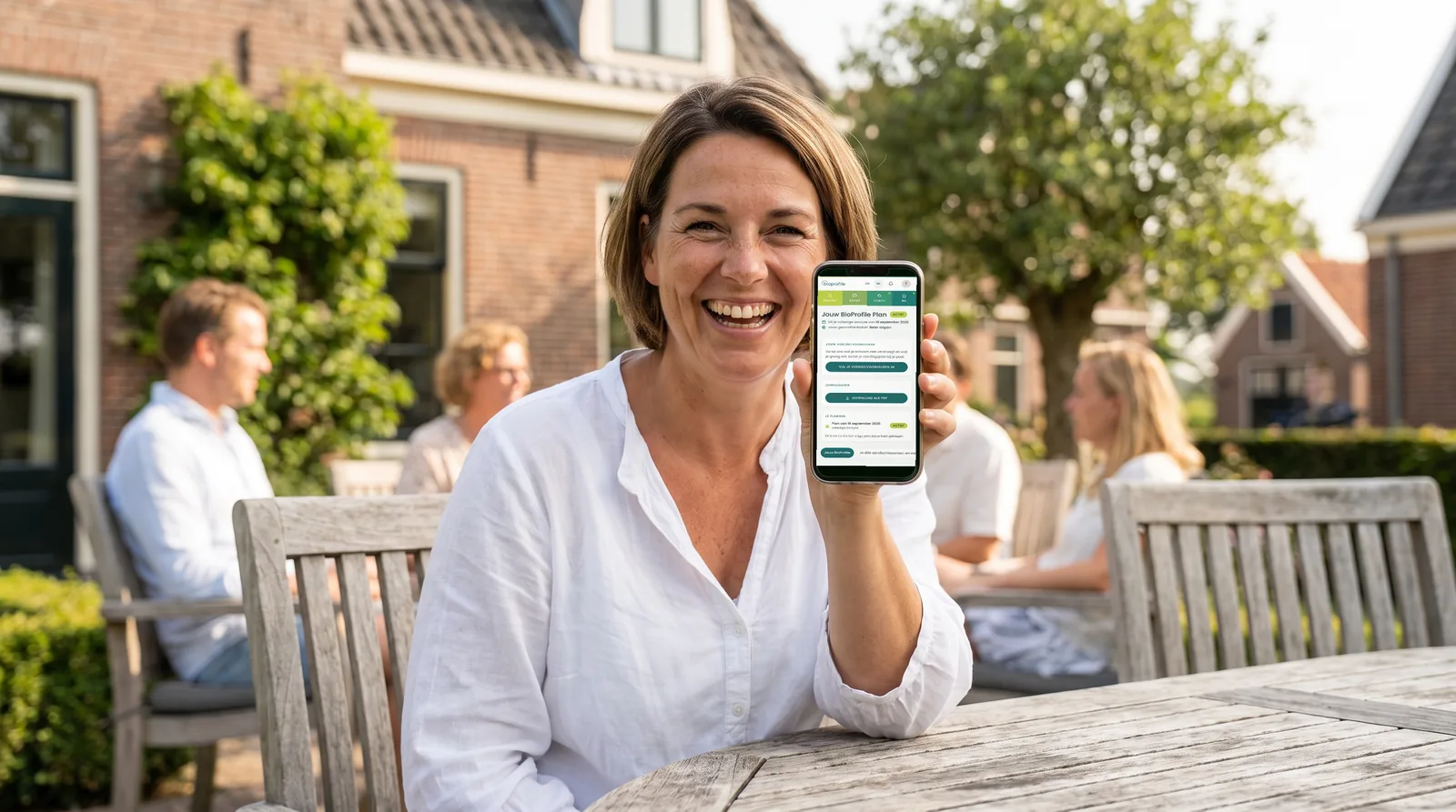 Een lachende vrouw van begin vijftig in een witte blouse zit op een terras in de ochtendzon en laat op een telefoon een BioProfile-plan in de app zien.