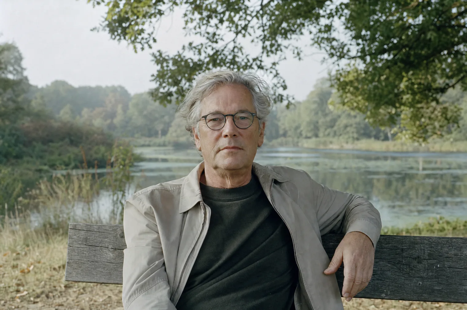 Ein Mann in den 60ern mit grauem Haar und Brille, in einer hellen Jacke, sitzt auf einer Holzbank an einem Teich, einen Arm über der Lehne, und blickt geradeaus.