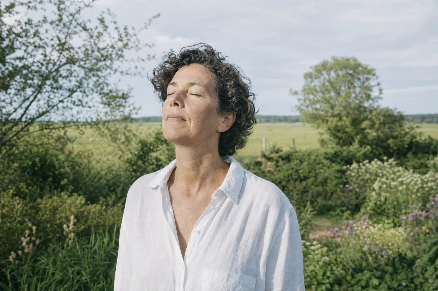 Eine Frau um die 50 mit kurzem, dunklem, lockigem Haar und weißem Leinenhemd steht mit geschlossenen Augen und dem Gesicht zur Sonne in einem grünen Garten am Rand einer flachen Weide.