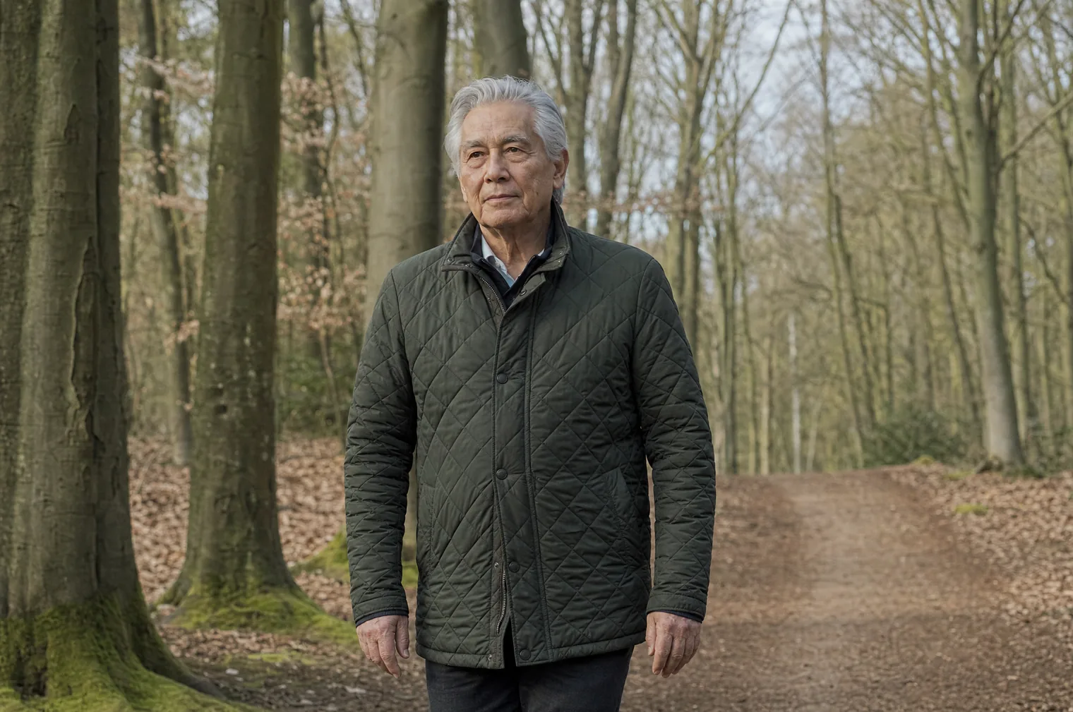 A man of around 70 with grey hair combed back and a dark green quilted jacket walks along a forest path between tall beech trees.