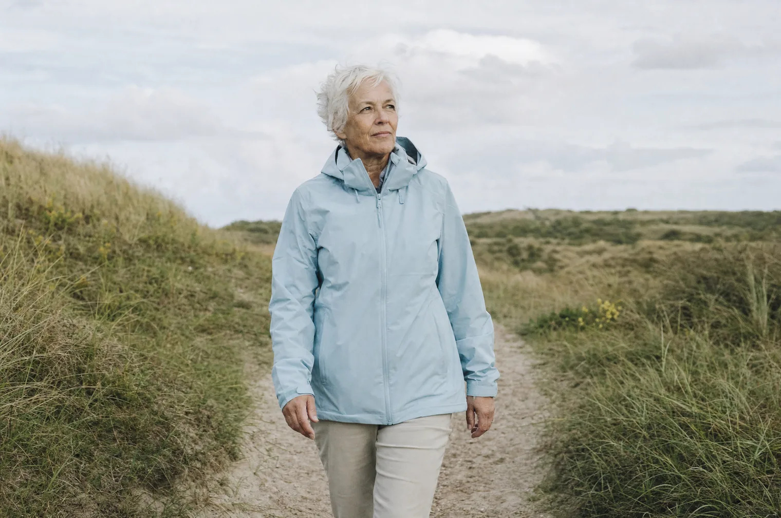 A woman aged 60-something, with short white hair and a light blue raincoat, walks along a narrow sandy path between overgrown dunes.
