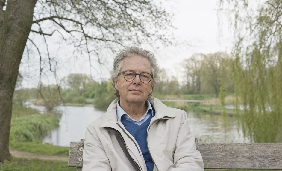Ein Mann Anfang 60 mit grauem Haar und Brille, in einer hellen Jacke über einem blauen Pullover, sitzt auf einer Holzbank an einem Teich mit einer Trauerweide und blickt geradeaus.