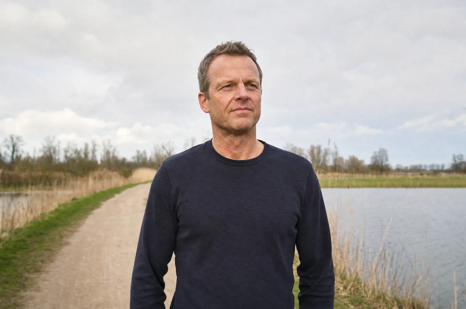 A man of around 50 with short blond hair and a dark jumper stands on a path beside a lake with reeds, looking calmly to one side.