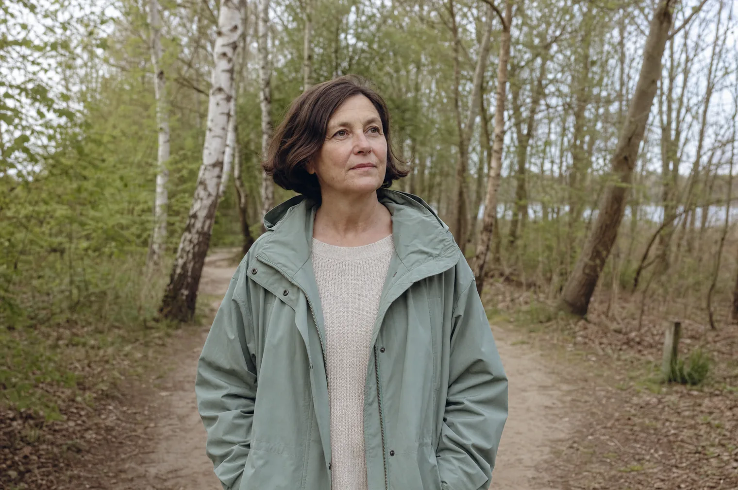 A woman of around 55 with dark hair in a bob and a sage green coat stands on a woodland path among birches.