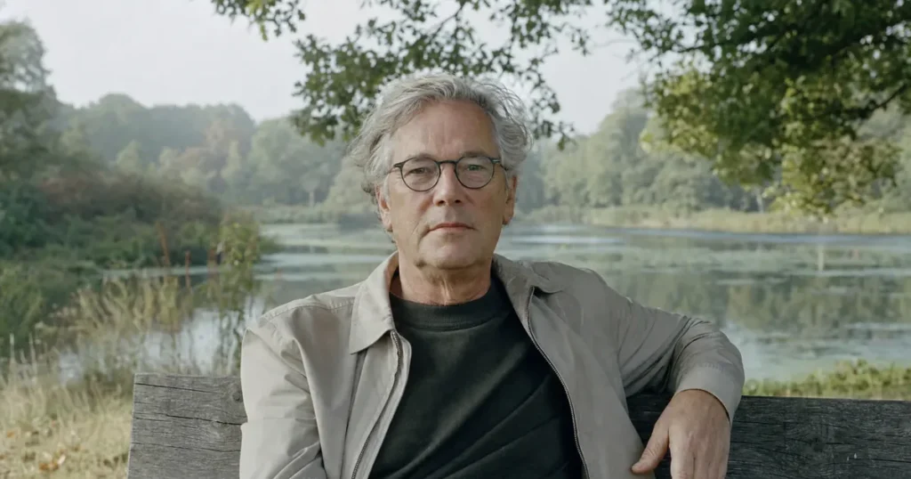 Een man van in de 60 met een bril en een lichte jas zit rustig op een houten bank bij een vijver, met een arm over de leuning, en kijkt recht vooruit; foto bij een pagina over lage hartslag symptomen.