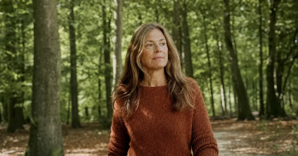 Een vrouw van rond de 50 met lange lichtbruine lokken en een roestbruine trui wandelt over een pad door een zonnig beukenbos, bij de vraag welke vitamine bij vermoeidheid helpt.